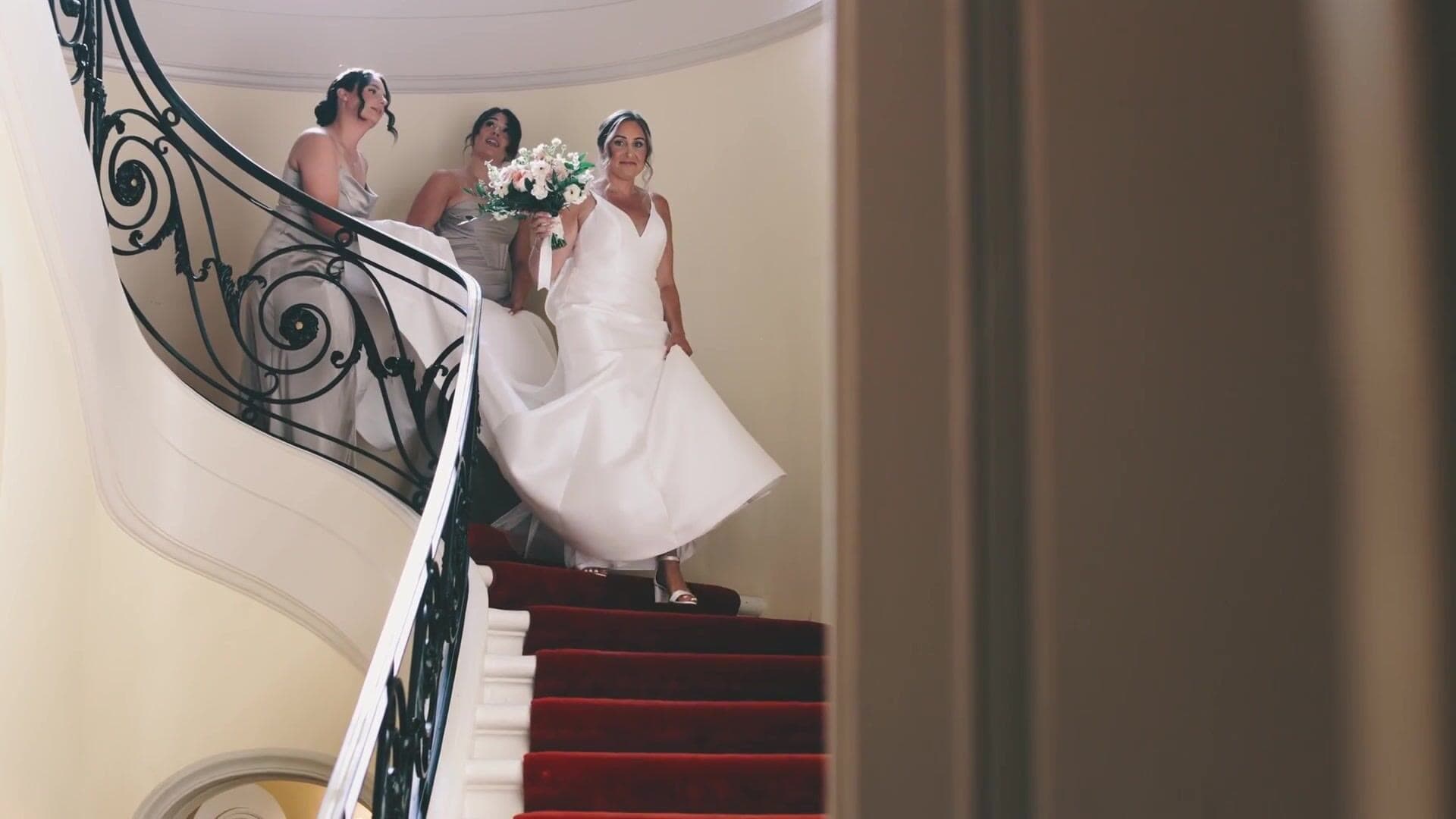 A bride coming down the curved staircase with a wrought-iron balustrade at Glen Manor House, her bridesmaids behind her