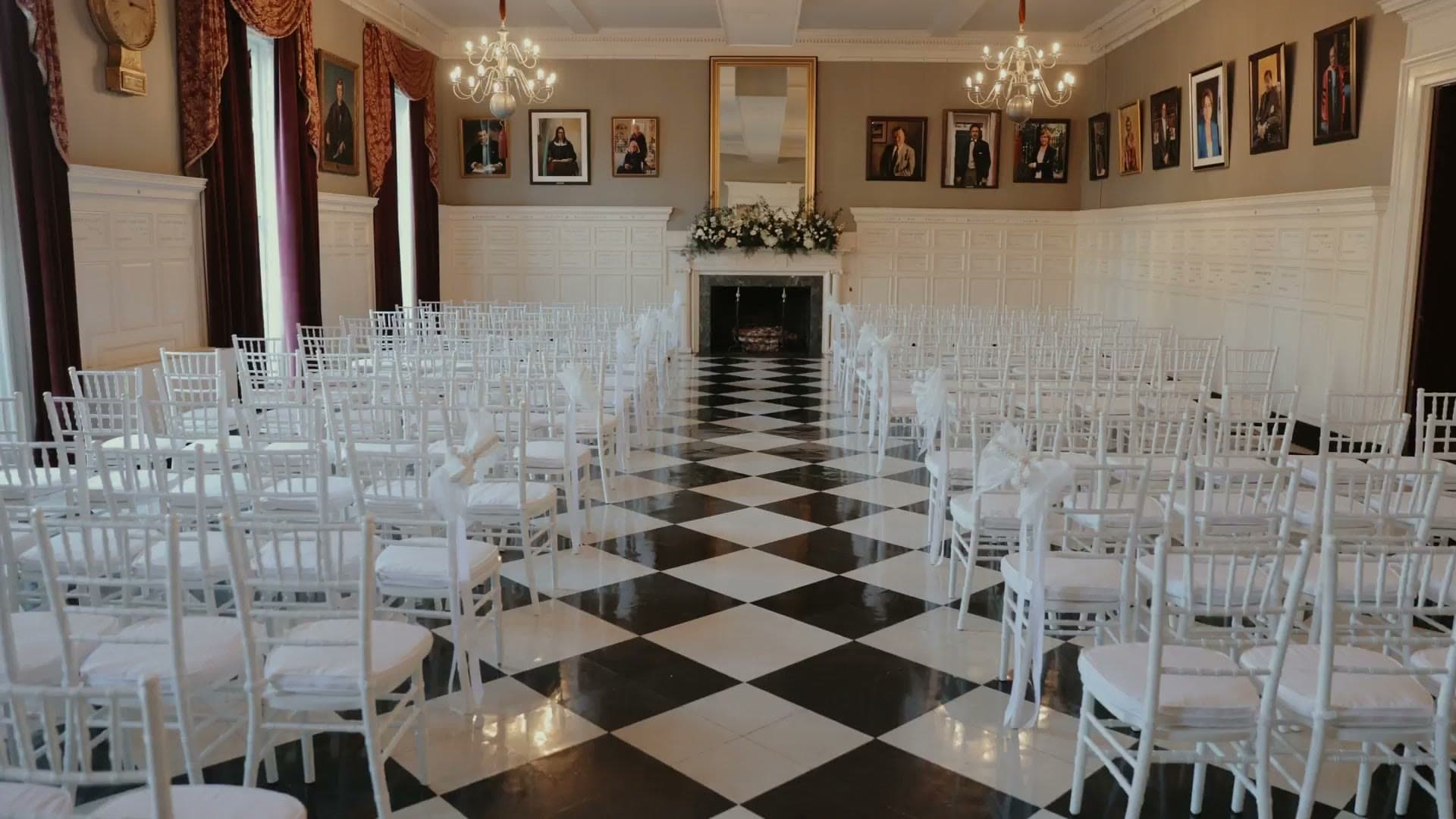 A white-panelled room at the Harvard Club of Boston set for a wedding ceremony, with portraits on the walls, chandeliers and a black-and-white checkerboard floor