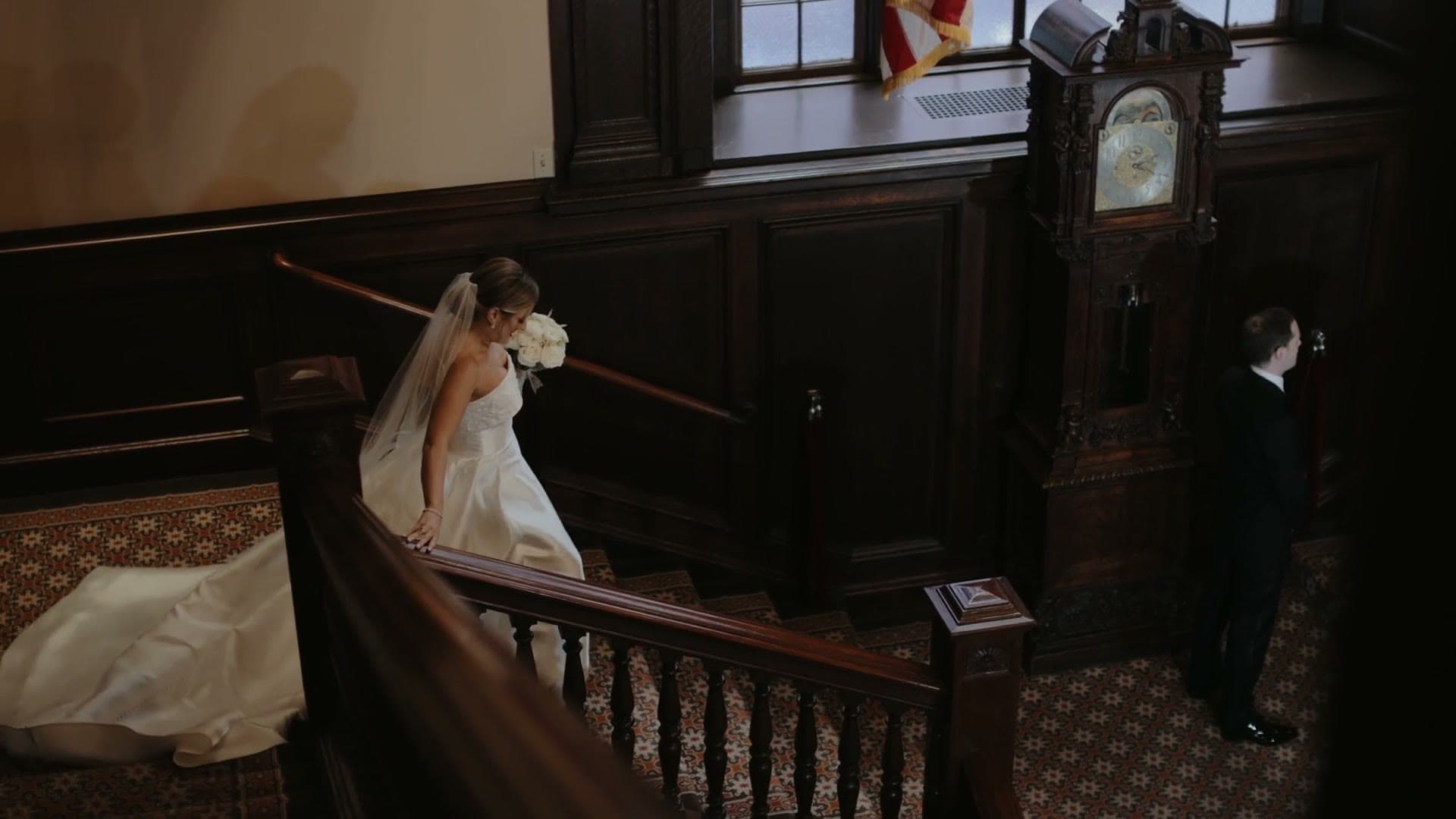A bride coming down the carved wooden staircase at the Harvard Club of Boston towards her groom, who waits beside a grandfather clock