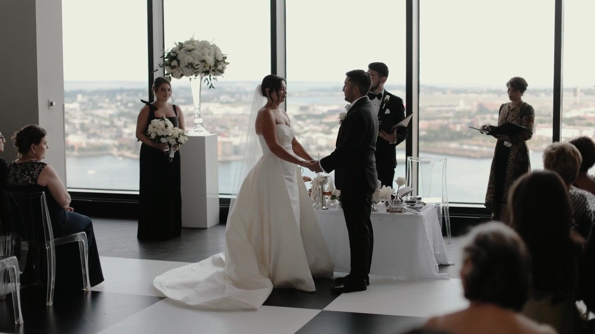 A bride and groom holding hands at their ceremony in front of floor-to-ceiling windows over Boston Harbor at the State Room, on a black-and-white checkerboard floor