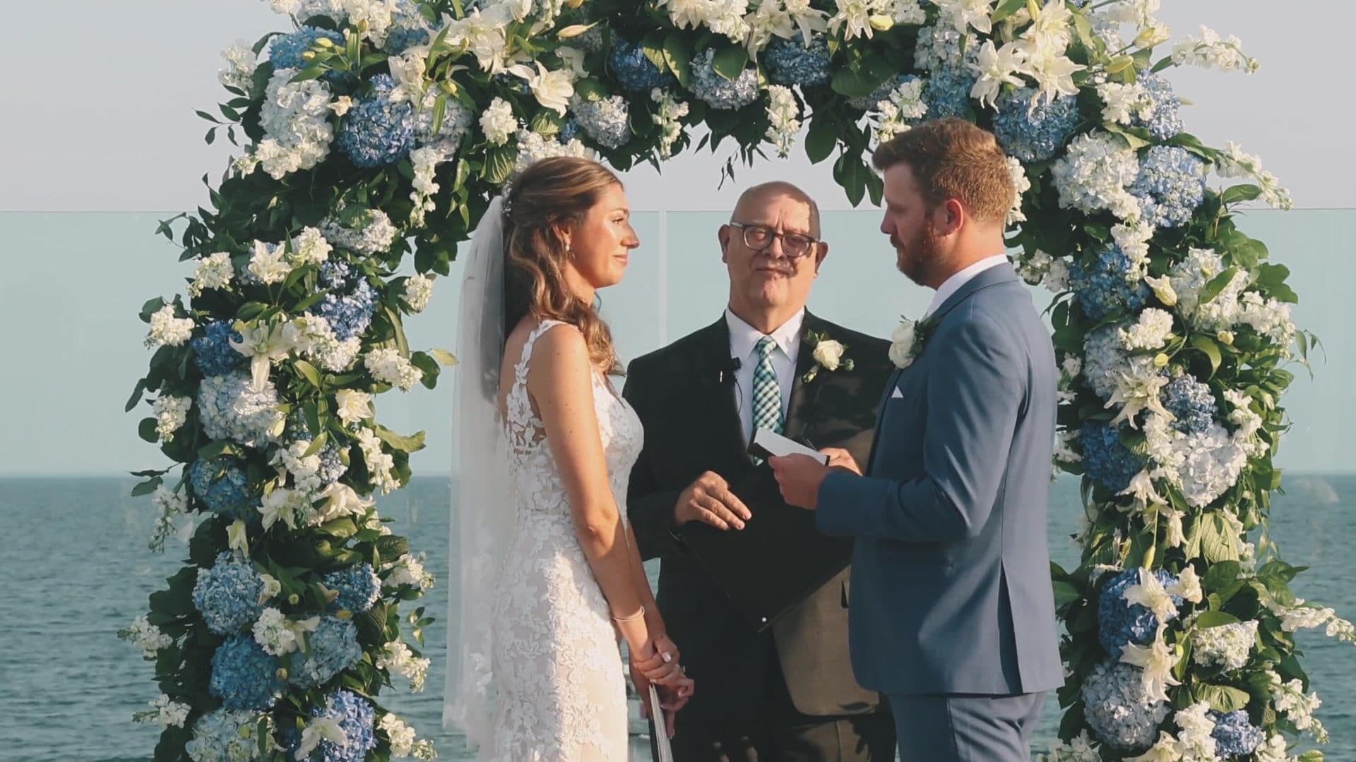 A couple exchanging vows beneath a blue and white hydrangea arch with Nantucket Sound behind them at Wychmere Beach Club