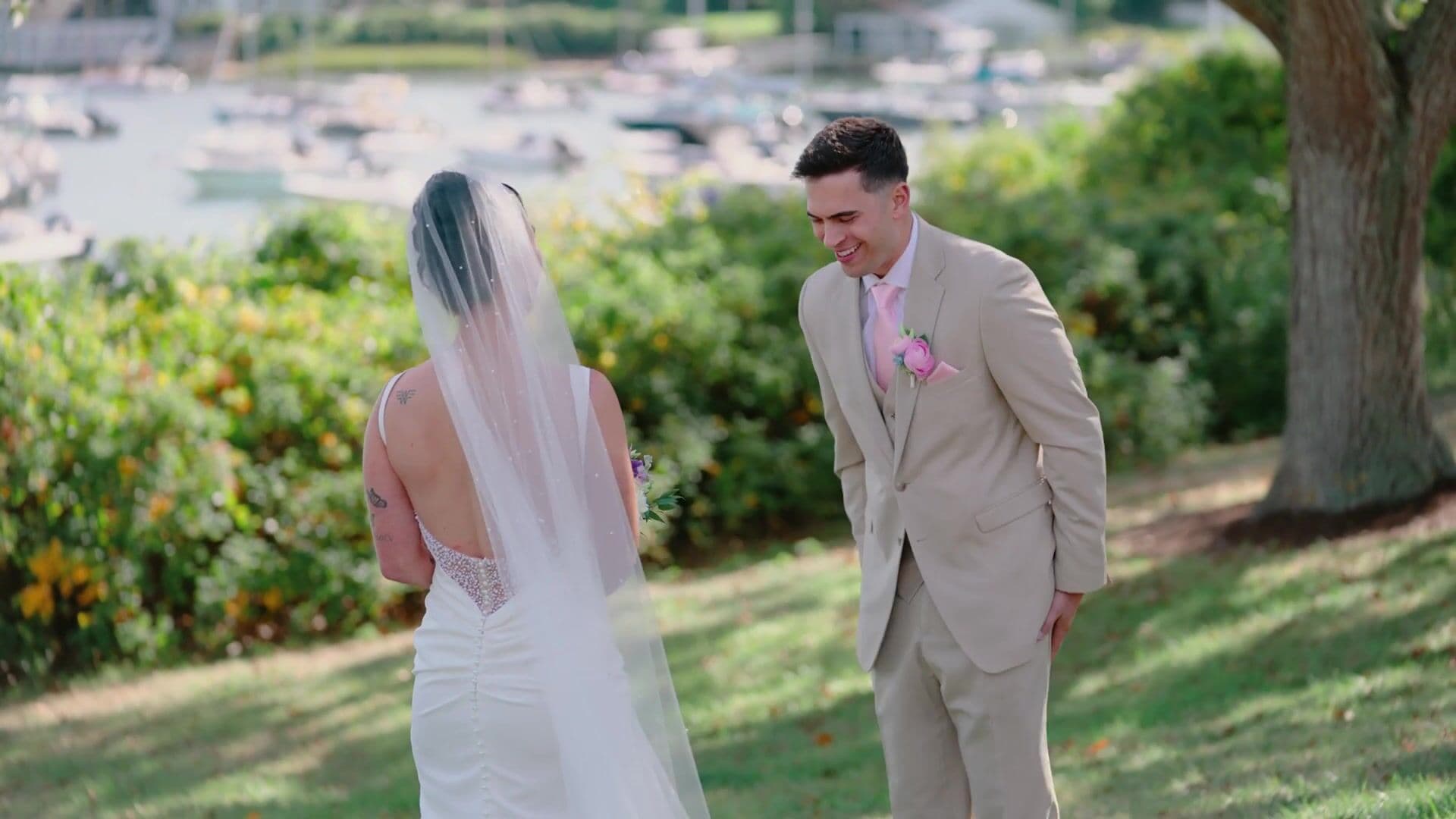 A groom turning to see his bride for the first time on a lawn above Wychmere Harbor, with moored boats behind them